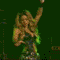 A woman with long wavy brown hair performs on stage in a dark green crop top and silver skirt, raising her arms against a green background.