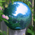 Pixelated image of a blue and green spherical object with a bright white reflection, set against a blurred green background with hints of pink and gray elements.