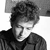 Black-and-white pixelated image of a person with curly hair wearing a dark jacket, head slightly tilted downward, expression neutral, background plain white.