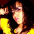 Pixelated close-up of a person with long brown hair wearing yellow clothing, holding an object near their face against dark background.