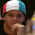A pixelated close-up of a person wearing a red, white, and blue cap with hand near chin against dark background.