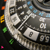 A circular analog dial with black, white, and metallic silver elements, featuring numbered markers and colored indicators around its perimeter.
