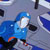 A pixelated blue figure with white highlights and dark accents appears against a gray background with scattered blue and black shapes.