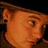 Pixelated close-up of a person's face with light brown hair, visible ear, and dark background, rendered in warm orange-brown tones.