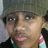 A pixelated close-up of a person with light skin tone wearing dark clothing and pink earrings, set against a blurred background.