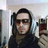 A person wearing dark sunglasses and a dark hooded garment holds a red digital camera in a room with light-colored walls.
