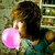 A person with brown hair blows a large pink bubblegum bubble while wearing a teal sleeveless top, standing in front of a blurred background.