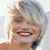 Pixelated close-up of a smiling person with light-colored hair, visible teeth, and a dark earpiece or accessory on the right side.