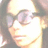 Pixelated close-up of a person with light skin tone wearing dark-framed glasses and long brown hair, smiling slightly against a bright background.