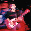 A person with light hair plays a red electric guitar on stage under dim lighting, captured in two showing slight movement