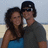 Two people stand close together on a beach, one in blue top and dark hair, the other in gray shirt and black cap.