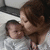 A woman with brown hair holds a baby in white clothing against a light background.