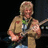 Pixelated image of a person with light hair wearing a beige shirt holding an electric guitar against dark background.