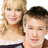 Two people with light and dark hair stand close together, the person on the left has long wavy blonde hair and wears a white top, while the person on the right has short brown hair.