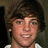Pixelated close-up of a person with dark brown hair, light skin tone, and visible teeth in a neutral expression against a muted background.