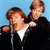 Two people in black jackets and white shirts stand before a microphone against a blue background, with one person's arm around the other's shoulder.