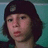 Pixelated close-up of a person with dark hair wearing a dark cap, set against a teal background.