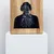 Pixelated static image of a dark figure in a light frame on a white pedestal.