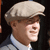 Pixelated close-up of a person wearing a light-colored flat cap and light blue shirt against dark background.