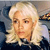 Static pixelated image of a person with light-colored hair wearing a dark top with a light collar, set against a blurred background with indistinct shapes.