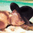 A person in a black cowboy hat and sleeveless top appears lying on sand, then standing upright against a beach backdrop with green water.