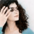 A pixelated close-up of a person with dark wavy hair, light skin, and a hand raised near their face against a light blue background.