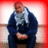 Pixelated figure in dark blue jacket with light gray patterned scarf, seated against red and white gradient background.