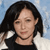 Pixelated image of a person with dark shoulder-length hair wearing a dark top against a light blue background with white cloud-like shapes.