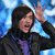 A person with long dark hair wearing a black jacket over purple clothing, raising their right hand in front of a blue pixelated background.