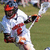 A person in a white uniform with red accents and an orange helmet holds a lacrosse stick on a grassy field.