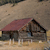 Static pixelated image of a small structure with a reddish-brown roof and dark walls on a light brown ground, set against a muted background.