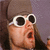 Pixelated close-up of a person wearing white sunglasses with black lenses, brown hair, and an open mouth expression against a purple background.