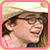 Pixelated static image of a person with long brown hair wearing glasses and a pink top, framed by a pink border on a light background.