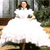 A pixelated image shows a person in a white dress standing outdoors with greenery and a building in the background.