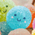 A pixelated blue circular icon with a smiling face, pink cheeks, and dark eyes appears in the foreground of a colorful background featuring green and orange shapes.