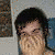 A pixelated image of a person with dark hair wearing blue jeans and a black belt, covering their face with both hands in front of a light-colored wall.