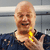 A pixelated image of a bald man with a brown beard wearing a dark blue shirt and a yellow necklace, holding a yellow object near his chest.