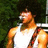 A pixelated image of a person in a white tank top with brown straps, standing against green foliage and metal railings.