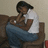 Pixelated image of a person sitting on a brown chair, wearing a white shirt and blue pants, with a light-colored wall background.