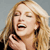 A pixelated close-up of a person with long blonde hair holding a black phone to their ear, mouth open in a shout or laugh against a gray background.