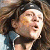 Two figures with long brown hair wear dark headbands and open mouths, positioned side-by-side against a blurred background.