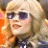 Pixelated close-up of a person with light-colored hair wearing dark sunglasses and a dark top against a blurred background with orange and brown tones.