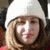 A person with light brown hair wears a white knit hat and red clothing, with a blurred background.