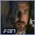 Pixelated image of a person with light skin and brown hair, wearing a dark jacket over a light shirt, standing against a dark background with visible text 'FAN' at bottom left.