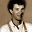 Static pixelated image of a person with dark hair wearing a white shirt and black necklace against a brown background.