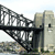 A pixelated static image shows a steel truss bridge with a dark horizontal beam, set against a light sky and distant greenish terrain.
