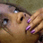 A pixelated close-up of a person's face with light skin, dark hair, and purple nail polish on visible fingers, set against a muted brown background.