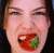 A pixelated close-up of a person's face with an open mouth holding a red and green object, set against dark hair.