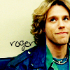 A person with light brown curly hair wears a blue jacket over a teal shirt and layered necklaces, with handwritten text 'roger' on a light background.
