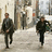 Three figures in dark clothing walk toward the viewer on a light-colored path with blurred background elements.